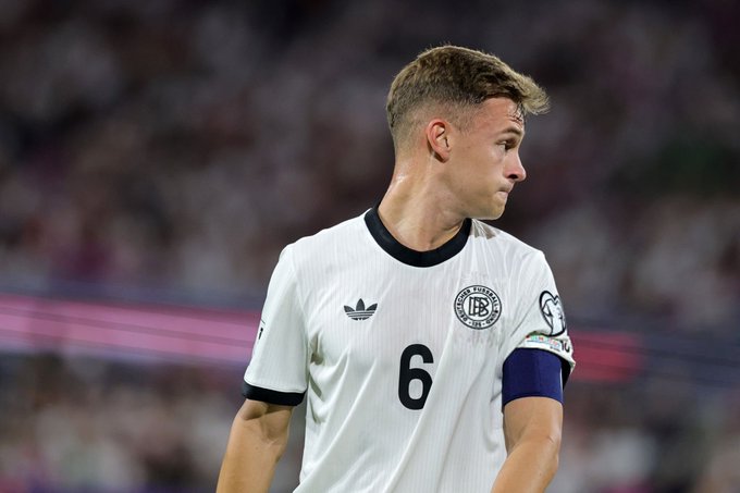 A young male footballer with short blonde hair stands on a stadium field wearing a white and black Adidas jersey with number 6 on the back and the number 6 patch on the sleeve the jersey features a circular emblem on the chest and navy blue accents on the collar and cuffs the background shows a blurred crowd in the stands.